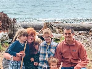 family of husband, wife, and four young children smiling in front of the ocean